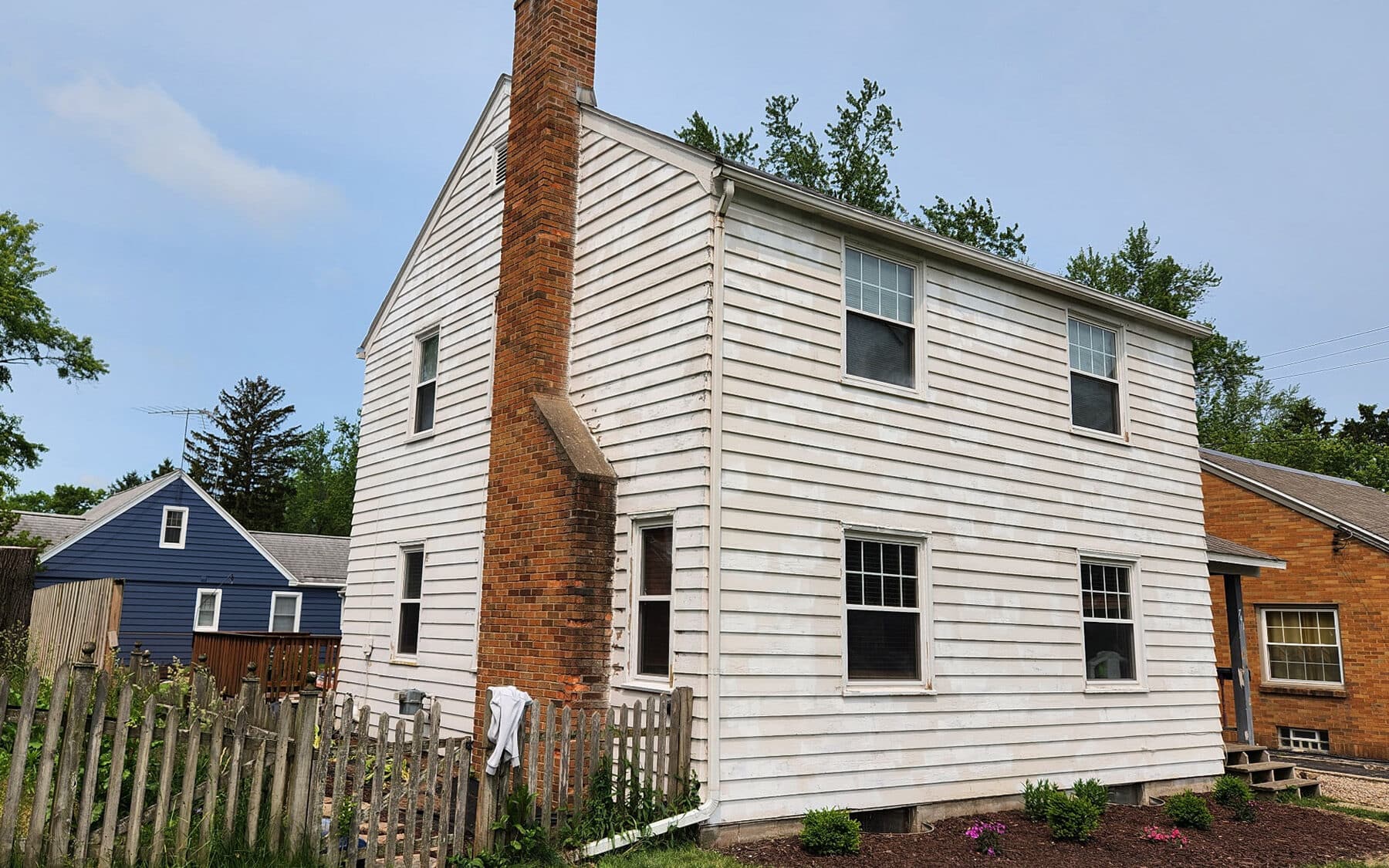Side view of two-story colonial home with weathered white siding and brick chimney before exterior repaint by Fortoso Painting in Kalamazoo, Michigan